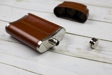 An open steel leather alcohol flask lies on a white wooden table. Portable outdoor kit with stacks...