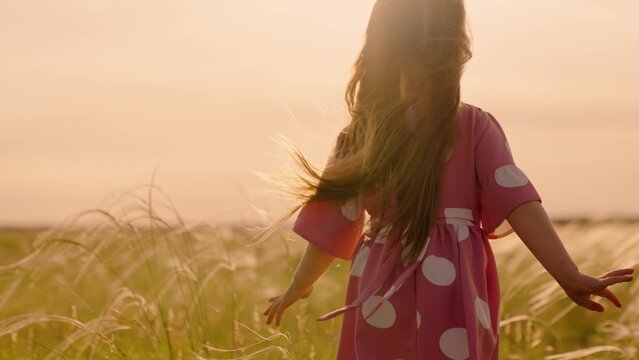 Joyful little girl in summer dress runs in field under rays of sunset. Happy child spends vacations in countryside. Little daughter plays in meadow, runs through tall green grass, child dream Family