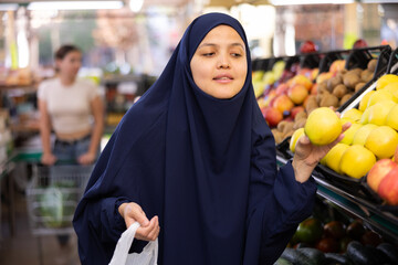 Young woman in paranja shopping and choosing local juicy apples in hypermarket