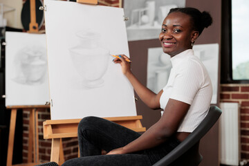 Happy woman sitting on chair at easel and smiling at camera, holding pencil creating masterpiece during group drawing class. Creative leisure activities and mental health concept