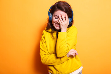 Fototapeta premium Cheerful lady covering face and listening to song on headphones in studio. Joyful woman enjoying song and sounds using headset while holding hand over closed eyes. Adult feeling happy.