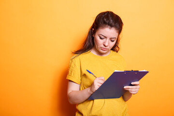 In the studio, young woman is using a pen to taking notes on paper. Focused individual signing project related paperwork on a notepad clipboard and writing in notebook files.