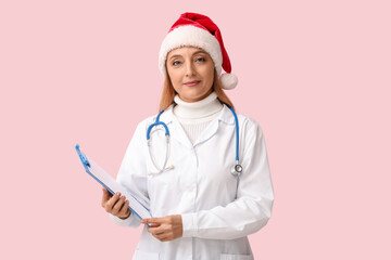 Mature female doctor in Santa hat with clipboard on pink background