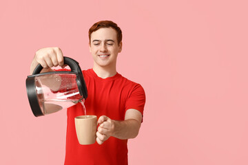 Handsome young man pouring hot water from modern electric kettle into cup on pink background