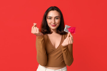 Young woman making heart shape with her fingers and holding condoms on red background. Safe sex concept - obrazy, fototapety, plakaty