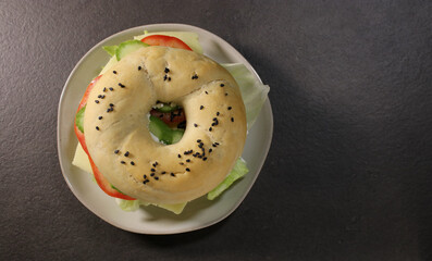 Top view of bagel with vegan cheese and fresh vegetables on a plate, dark grey table