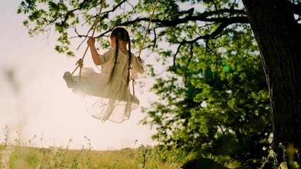 Child plays on wooden swing, dreams of flying. Baby swing, kid girl smile in flight. Happy little girl swings on swing in park under tree at sunset. Concept of family holiday, dreams, entertainment. - obrazy, fototapety, plakaty
