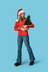 Beautiful young woman in Santa hat with Christmas tree on blue background