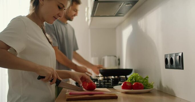 A brunette girl in a white T-shirt is preparing a morning salad and her husband, a guy in a gray T-shirt, talks to her and cooks something on the stove in the kitchen in the morning