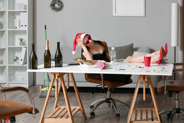 Drunk young woman sitting at table in office after New Year party - obrazy, fototapety, plakaty