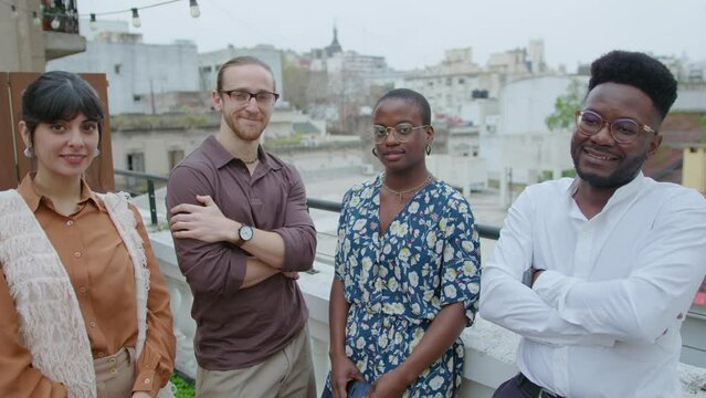 Group of young multi-ethnic colleagues posing for camera and smiling outdoors at urban rooftop. Video portrait, cityscape in the background