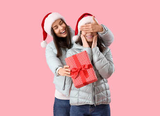 Young woman surprising her friend with Christmas gift on pink background
