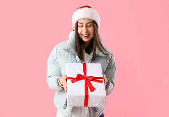 Young woman in Santa hat with Christmas gift on pink background