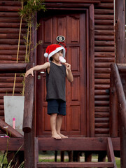 Latin little boy outside a wooden cabin, holding a popsicle in his hand, celebrating Christmas in summer. Wearing a red Santa Claus hat and sleeveless t-shirt. vertical. Argentina.