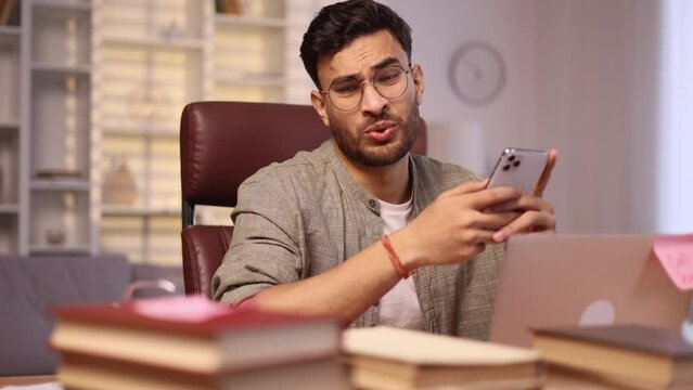 Boring exhausted young man student or freelancer scrolling feed of social networks products in internet store on smartphone instead of work or education at home workplace Procrastination concept