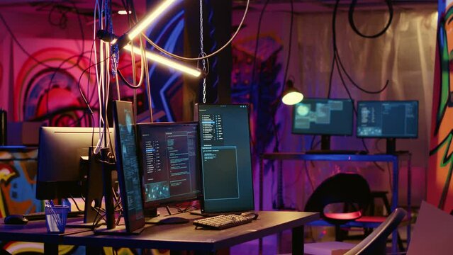Programming language on PC screens in empty messy hackers base of operations with grunge graffiti drawings sprayed on walls. Neon lit ghetto hideout used by criminals to commit illegal activities