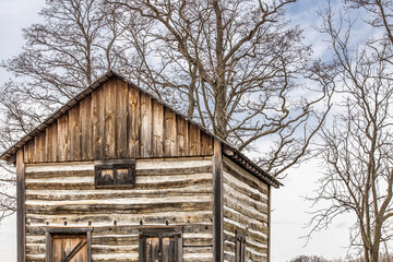 The upper story of a log cabin with leafless trees in the background in the autumn. 