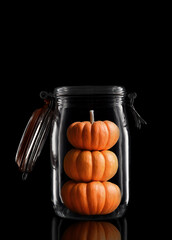 Three Decorative Mini Pumpkins stacked in a glass storage or canning jar isolated on black with reflection, with lid open.