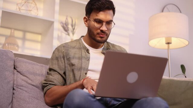 Handsome man with glasses browse products in internet online shopping store planning trip looking for job or swiping through profiles on dating service on laptop computer sitting on couch at home