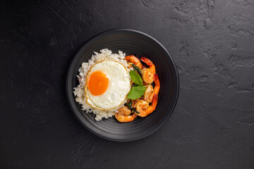 Stir-fried shrimp with basil Thai street food arranged on a black plate with a fried egg Thai spicy food Placed on a dark black table. Top view.
