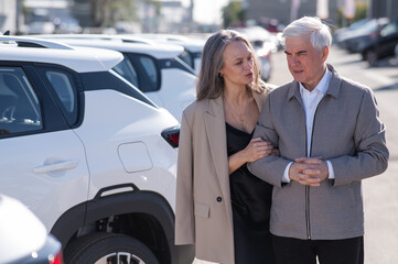 Mature Caucasian couple walks past cars outdoors, choosing a new one. 