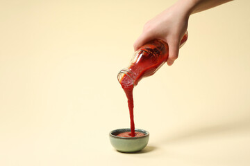 Woman pouring tasty ketchup from bottle into bowl on beige background, closeup. Space for text