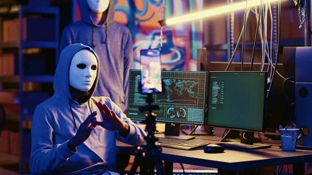 Masked hackers filming ransom video in secret HQ, threatening to release stolen data publicly if payout demands are not met. Cybercriminals using smartphone to record blackmail footage