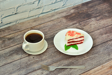 A cup of hot coffee on a saucer and a plate with a piece of red velvet cheesecake with mint leaves on a wooden table.