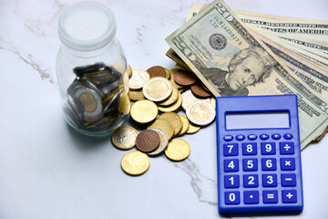 Glass jar with multicurrency coins, US dollar bills and calculator white table.