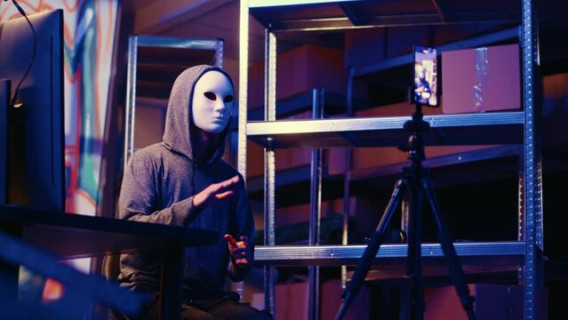 Hacker wearing anonymous mask doing ransom video after gathering information from users computers using spyware software. Cybercriminal blackmailing victim in graffiti painted hideout