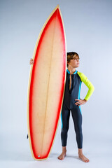 Young surfer boy wearing a wet suit posing beside his surfboard in a white studio 