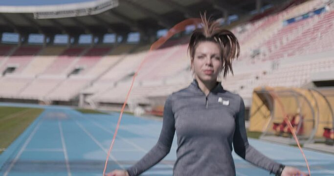 Female runner girl jumping with rope on synthetic sports track. Looking at camera. Out of focus open field generic stadium in the backgorund.
