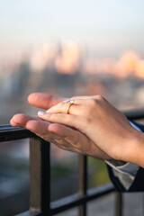 A man's hand holds a woman's hand to show off a diamond ring after proposing. The background is a pastel bokeh of the city skyline.