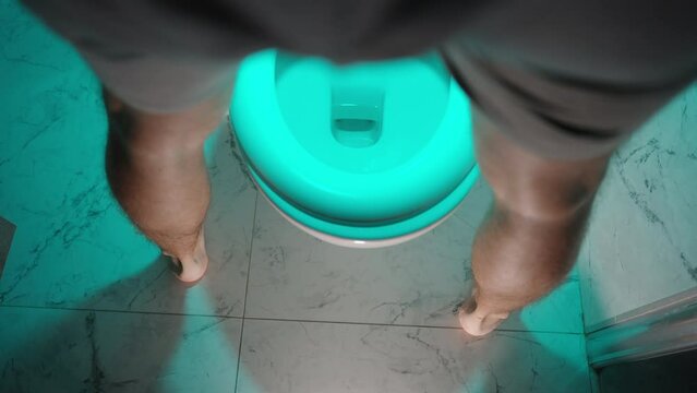 Top View of a Toilet in Blue Light at Night a Man in Shorts Stands in Front of It to Attend to His Needs.