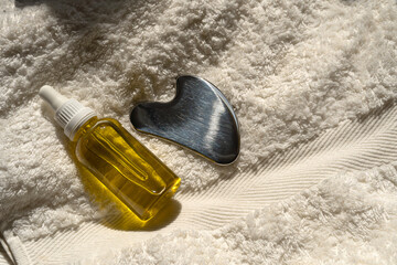 a bottle of oil and a stainless steel gua sha scraper lie on a towel