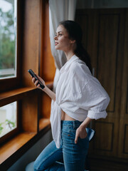 Young woman standing at the window with phone in hand smiling and looking out the window at nature,...