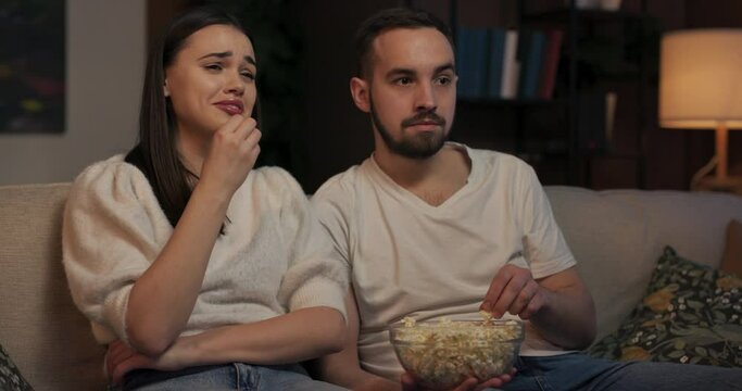 Couple sitting on couch in cozy living room munching on popcorn watching TV relaxed and content TV show playing in background drama sitcom occasionally chuckle at the jokes comment on plot.