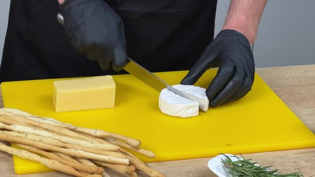 Person in black apron and gloves preparing cheese on a yellow cutting board with breadsticks and fresh herbs on the side