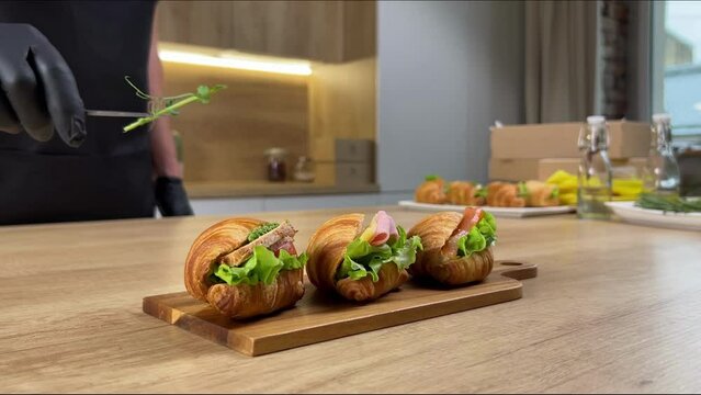 A chef's gloved hand garnishes freshly prepared croissant sandwiches on a wooden board in a modern kitchen setting