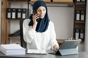 Happy muslim woman talking to smartphone sitting at desk in office.