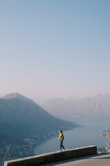Fototapeta na wymiar Young woman stands on a stone curb above the Bay of Kotor high in the mountains. Montenegro