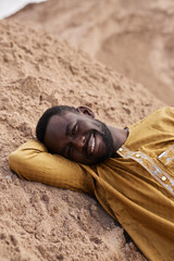 Vertical portrait of Black young man lying on sand dune in desert and smiling at camera