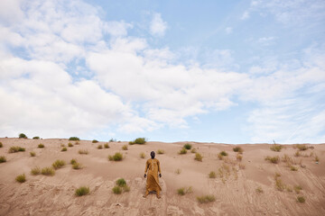 Dramatic wide angle shot of traditional Black man wearing long kaftan standing on sand dune in desert landscape with wind blowing, copy space