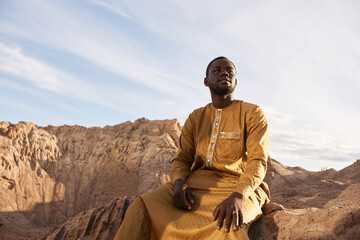 Dramatic portrait of Black young man wearing long kaftan sitting sand dune and looking at sky in...
