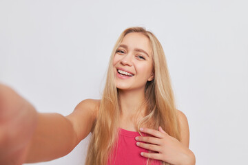 Fototapeta premium Portrait photo of happy blonde girl dressed in pink top doing selfie in white studio smiling, beauty concept, copy space