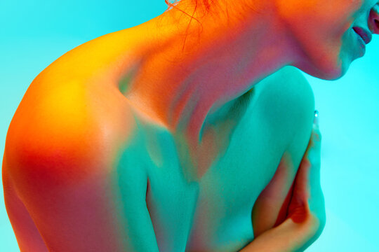 Cropped image of naked female body, woman sitting and covering breast with hand against blue background in neon light. Body aesthetics, beauty