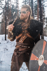 Fototapeta premium A fierce Viking warrior poised with an axe and shield in a snowy pine forest, depicting strength and historical Nordic culture