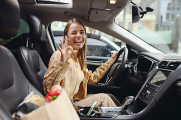 Young smiling happy customer woman wear casual clothes show ok driving car with paper bag of goods...