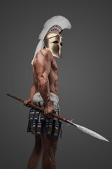 An impressive portrait of a bare-chested ancient Greek hoplite, wearing a helmet and brandishing a spear, standing in a studio with a gray backdrop