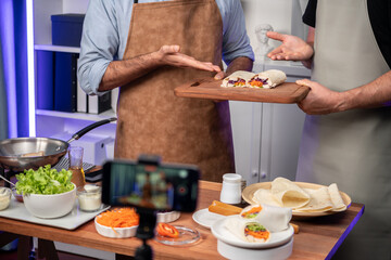 Cropped view of chef influencers presenting fresh salad roll on cooking step by step, streaming via smartphone on social media live channel, showing ingredients vegetable soft tortilla wrap. Sellable.